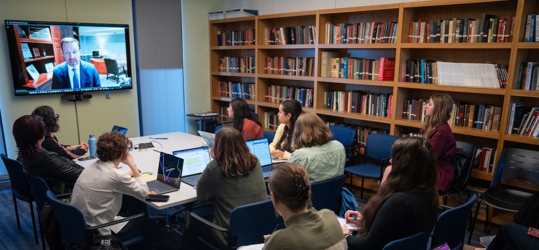 College students in a library classroom watch and listen to a man on a screen.